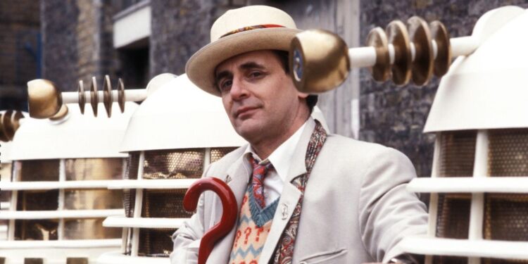 Sylvestor McCoy in costume as the Seventh Doctor surrounded by white Daleks