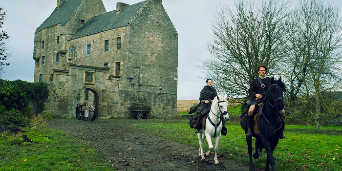 Two individuals riding a horse with a castle behind them in Outlander